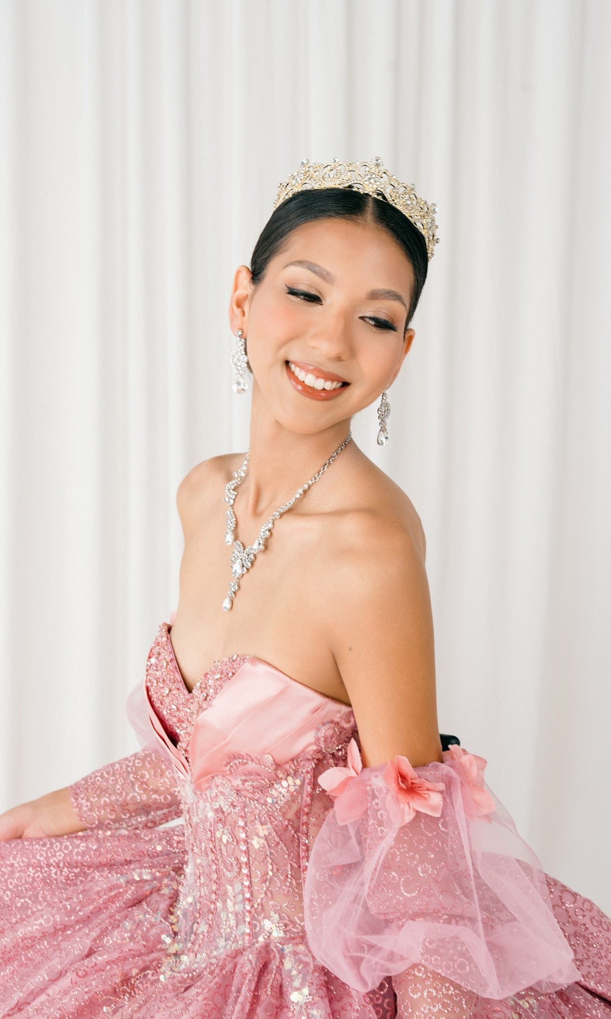 Woman wearing the Quinceanera Dress 8385J by Cinderella Couture, featuring a sparkling pink corset bodice and jeweled tiara, smiles while looking to the side in front of a white curtain backdrop.